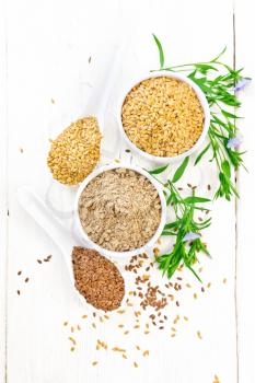 Flaxseed flour in a bowl, white and brown linen seeds in two spoons and on table, leaves and blue flax flowers on background of light wooden board from above