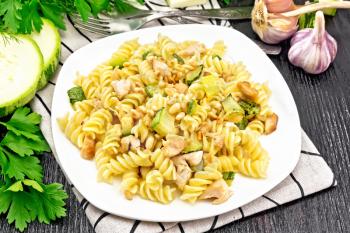 Fusilli pasta with chicken breast, zucchini, cream and pine nuts in a plate on towel, garlic, fork and parsley against a dark wooden board