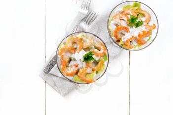 Puff salad with shrimp, avocado, fresh cucumber, sweet pepper and tomato, seasoned with yogurt sauce in two glass glasses on a napkin, bread and forks on a wooden board background from above