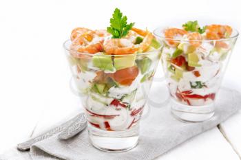 Puff salad with shrimp, avocado, fresh cucumber, sweet pepper and tomato, seasoned with yogurt sauce in two glass glasses on a napkin, bread and forks on a white wooden board background