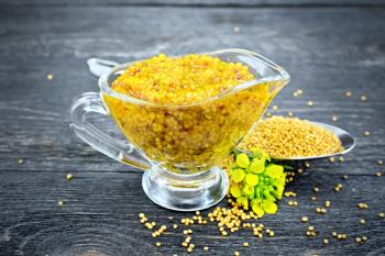 Mustard Dijon sauce in a glass sauceboat, yellow flowers and mustard seeds in a spoon on a background of a dark wooden board