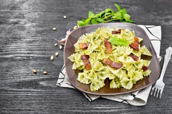 Farfalle pasta with pesto sauce, fried bacon and basil in a plate on a towel on wooden board background