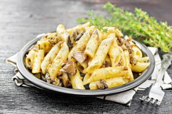 Penne pasta with wild mushrooms in a plate on a kitchen towel, thyme, fork and garlic on black wooden board background