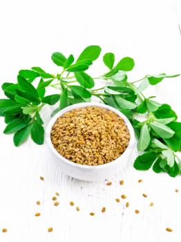 Fenugreek seeds in a bowl and on a table with green leaves on background of light wooden board