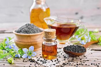 Nigella sativa oil in vial, gravy boat and bottle, seeds in a spoon and black cumin flour in a bowl on a burlap napkin, kalingi twigs with blue flowers and leaves on wooden board background