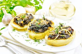 Potatoes stuffed with mushrooms, fried onions and cheese in a plate on towel, vegetable oil in carafe, parsley, garlic and a fork on background of white wooden board