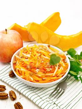 Pumpkin, carrot and apple salad with pecans seasoned with vegetable oil in a bowl on a napkin, mint on white wooden board background