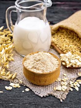 Flour oat in bowl, milk in jug, oatmeal in spoon on burlap, grain in bag, oaten stalks against dark wooden board