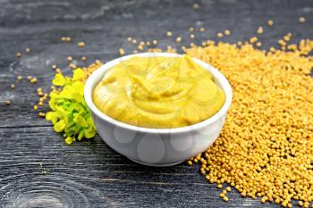Mustard sauce in a white bowl, mustard flower and seeds on a wooden board background