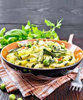 Tagliatelle pasta with zucchini, green peas, asparagus beans, hot peppers and spinach in a plate on brown towel, garlic, fork and basil on wooden board background