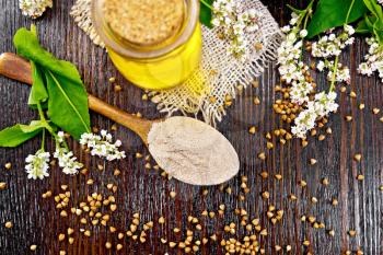 Buckwheat flour from brown cereals in a spoon, oil in a glass jar on sacking, flowers and leaves on the background of wooden board from above