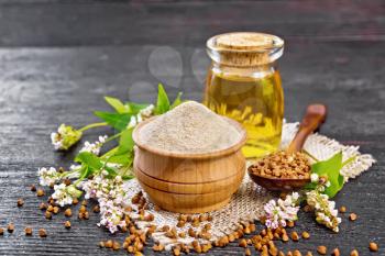 Brown buckwheat flour in a bowl, brown groats in a spoon, buckwheat flowers and leaves, oil in a glass jar on a burlap napkin on dark wooden board background