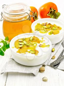 Yogurt, persimmon and honey dessert with vanilla, cardamom and pistachios in two bowls on a napkin, mint and spoon on wooden board background