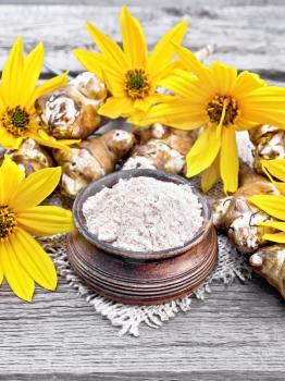 Jerusalem artichoke flour in a clay bowl on a burlap with yellow flowers and vegetables on background of an old wooden board