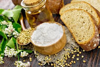 Buckwheat flour from green cereals in a bowl on sacking, groats in spoon and on a table, oil in a glass jar, bread, fresh flowers and buckwheat leaves on wooden board background