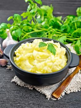 Mashed potatoes in a black saucepan and a spoon on burlap, garlic, parsley on dark wooden board background