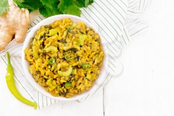 Indian national dish kichari of mung bean, rice, stalk celery, spinach, hot pepper and spices in a bowl on a napkin, ginger on wooden board background from above
