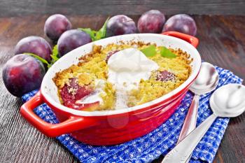 Crumble with plum and ice cream in a red brazier with spoons on a napkin, plums and mint on dark wooden board background