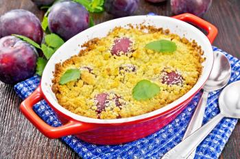 Crumble with plum in a red brazier with spoons on a napkin, plums and mint on dark wooden board background