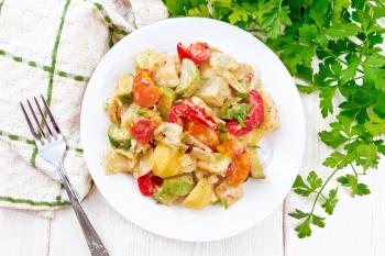 Vegetable ragout with zucchini, cabbage, potatoes, tomatoes and bell peppers in creamy sauce in plate, napkin, parsley and a fork on the background of light wooden board from above