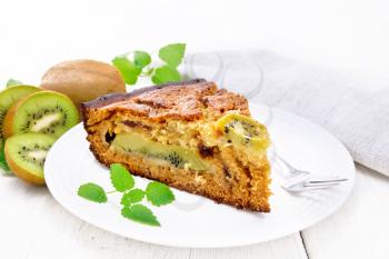 Piece of sweet cake with kiwi and honey, mint and a fork in a plate, a napkin on a light wooden board background