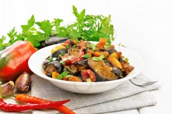 Vegetable ragout with eggplant, tomatoes, sweet and hot peppers, onions, carrots, fried with herbs and spices in a plate on napkin, garlic, parsley on wooden board background