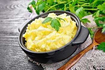 Mashed potatoes in a black saucepan and a spoon on burlap, garlic, parsley on black wooden board background