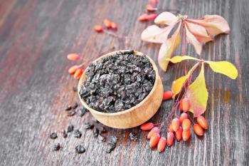 Dried black barberry in a bowl, sprigs with fresh berries and leaves on dark brown wooden board background