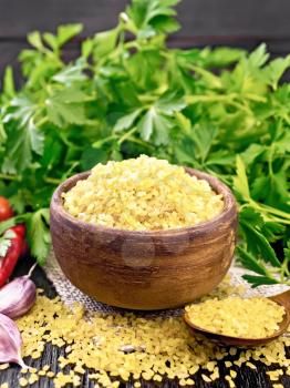 Bulgur groats - steamed wheat grains - in a clay bowl and a spoon on sacking, tomatoes, hot peppers, garlic and parsley on dark wooden board background