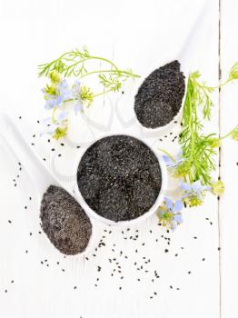 Black cumin seeds in a bowl, flour and seeds in spoons, kalingi sprigs with blue flowers and leaves on a light wooden board background from above