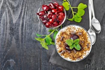Dessert Black forest of cherries, chocolate biscuit and soft cottage cheese with cream in a glass on a napkin, mint, berries in a bowl and spoon on wooden board background from above