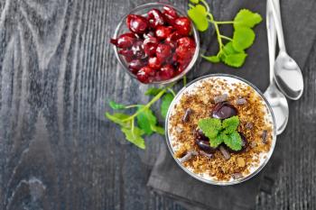 Dessert Black forest of cherries, chocolate biscuit and soft cottage cheese with cream in a glass on a napkin, mint, berries in a bowl and spoon on wooden board background from above