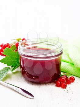 Red currant jam in a glass jar, bunches of berries with leaves, a towel and a spoon on the background of light wooden board