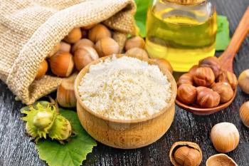 Hazelnut flour in a bowl, nuts in bag, a spoon, oil in glass jar and filbert branch with green leaves on wooden board background