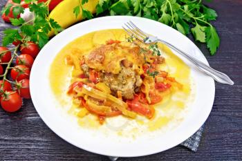 Chicken stewed with tomatoes, yellow and red bell peppers and cheese in white plate on a towel, thyme, parsley and garlic on wooden board background