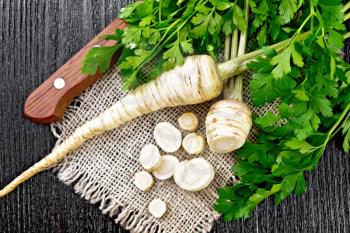Parsley roots whole and chopped with green tops on a burlap napkin and a knife on wooden board background from above