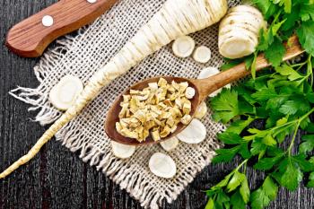 Fresh whole and chopped parsley roots with green tops, dried root in a spoon on burlap napkin, a knife on wooden board background from above