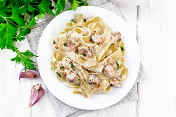 Tagliatelle pasta with salmon, cream, garlic and herbs in a plate on a napkin, fork, parsley and basil on the background of light wooden board from above