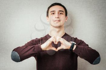 Closeup portrait handsome smiling young business man makes the heart using fingers, hands, isolated grey wall background. Positive human emotions, facial expressions, feelings, body language, attitude