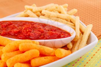 Potato, corn chips in bowl and red sauce on colorful tablecloth.