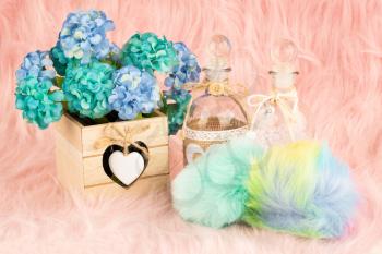 Beige wooden box, flowers, bottles and fluffy pompons on pink fur background.