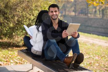 Beautiful Young Couple Sitting In The Park On A Beautiful Autumn Day - They Are Using Digital Tablet And Book