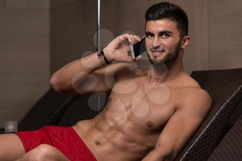 Young Man Resting On Sun Loungers By Swimming Pool And Talking On Mobile Phone