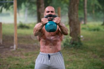 Strong And Muscular Young Fitness Man -  Holds Up A Green Kettlebell Outdoors  - Sports And Fitness - Concept Of Healthy Lifestyle - Fitness Male