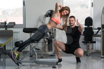 Personal Trainer Showing Ok Sign To Client - Young Woman Exercising Her Back On Machine In The Gym