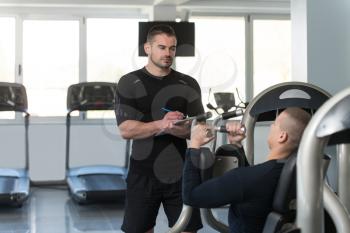 Personal Trainer Takes Notes While Young Man Exercise Chest On Machine In The Gym