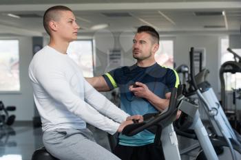 Young Man In The Gym with Personal Trainer - Exercising His Legs Doing Cardio Training On Bicycle