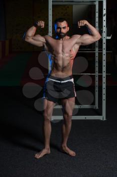 Young Man Standing Strong In The Gym And Performing Front Double Biceps Pose - Muscular Athletic Bodybuilder Fitness Model Posing Exercises