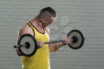 Muscular Man Doing Heavy Weight Exercise For Biceps With Barbell In Gym On White Bricks Background With Copyspace