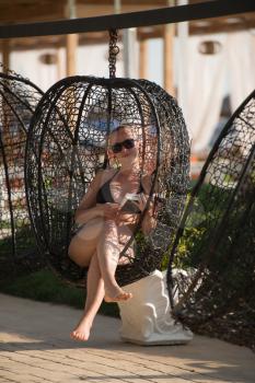 Young Woman Sitting in a Metal Rattan Hanging Basket With Cushions - Enjoying the Sunny Day in an Outdoor Reading a Book by the Sea